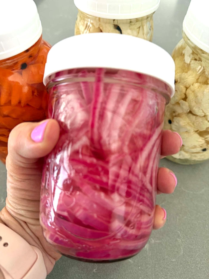 A small jar of red pickled onions with a white lid held in a hand with a jar of pickled carrots and two jars of pickled cauliflower behind it.