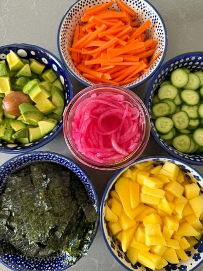 A small bowl of diced avocado, a small bowl of diced mango, a small bowl of matchstick carrots, a small bowl of slice cumbers, a small bowl of pickled red onions, and a small bowl of cut seaweed sheets.