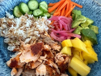 A bowl filled with blackened salmon flakes, rice drizzled with Japanese sauce, sliced cucumbers, matchstick carrots, diced avocados, pickled red onions, and diced mango.