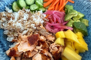 A bowl filled with blackened salmon flakes, rice drizzled with Japanese sauce, sliced cucumbers, matchstick carrots, diced avocados, pickled red onions, and diced mango.