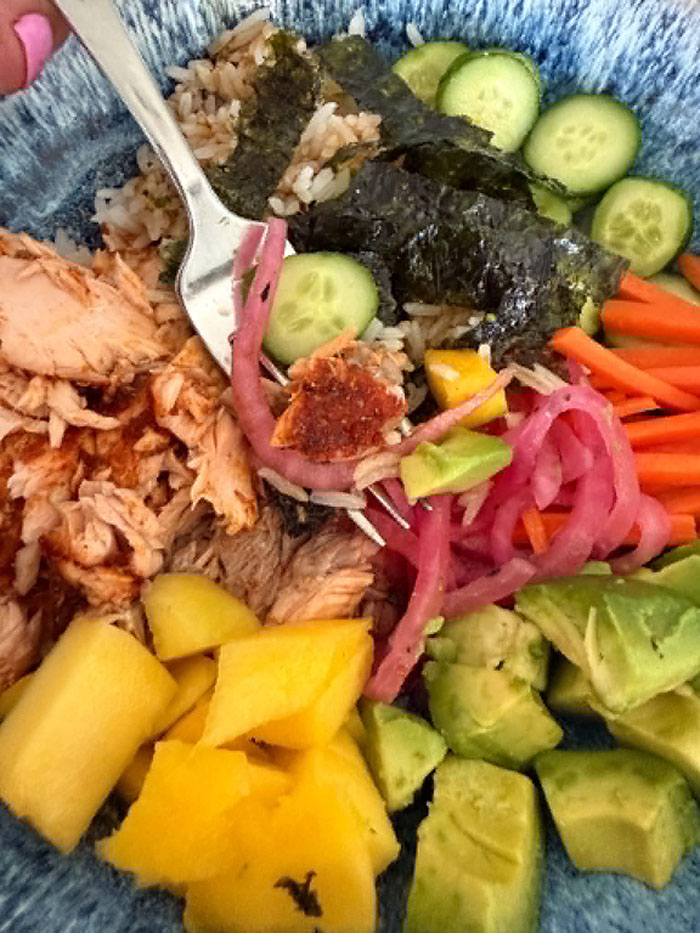 A bowl filed with diced avocado, matchstick carrots, sliced cucumbers, seaweed, rice with Japanese bbq sauce, flaked salmon, and diced mango. A fork is held diagonally from the top left sitting on top of the food with a bite of the food on it.