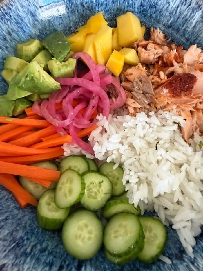 A bowl filled with sliced cucumbers, matchstick carrots, diced avocado, pickled red onion, diced mango, blackened salmon flakes, and white rice.