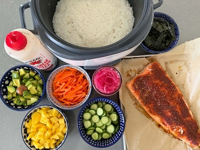 A small bowl of cut seaweed sheets, a rice cooker pot filled with rice, a bottle of Japanese barbecue sauce, a small bowl of diced avocado, a small bowl of diced mango, a small bowl of matchstick carrots, a small bowl of slice cumbers, a small bowl of pickled red onions next to a baking sheet with parchment paper and a blackened salmon filet on it.