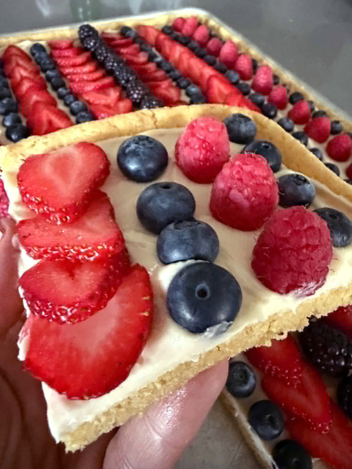 A corner slice of fruit pizza with strawberries, blueberries, and raspberries held above a Sheet Pan Fruit Pizza.