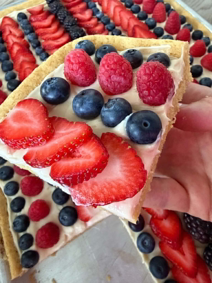 A slice of fruit pizza with blueberries, raspberries, and strawberries held above a Sheet Pan Fruit Pizza with rows of blueberries, raspberries, strawberries, and blackberries with a piece cut out.