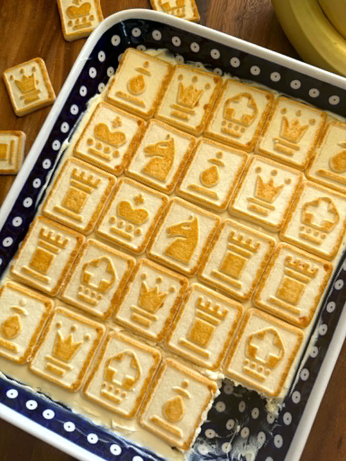 A dark blue and white polka-dotted 9x13 baking dish of Banana Pudding with Chessmen Cookies with a slice cut out of the bottom right corner. Four Chessmen cookies surround the top left corner of the dish. In the upper right corner is a partial picture of a banana.