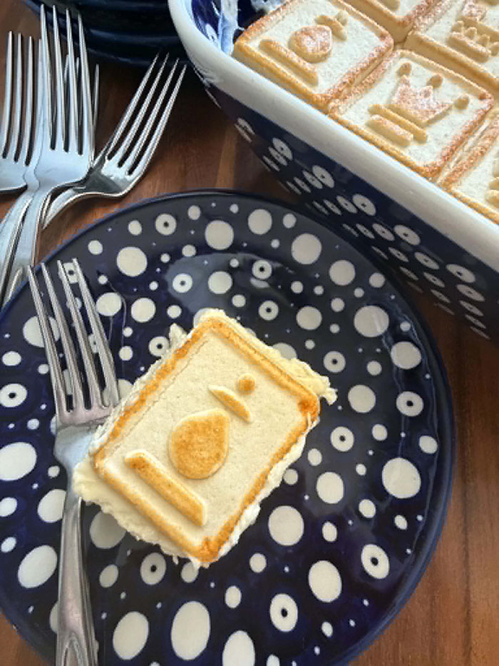 A slice of Chessmen Cookie Banana Pudding on a small dark blue plate with white polka dots. A fork is lying on the plate to the left of the slice of banana pudding. In the upper left corner are several forks stacked on each other. In the upper right corner is the corner of a 9x13 baking dish of Banana Pudding with Chessmen Cookies.