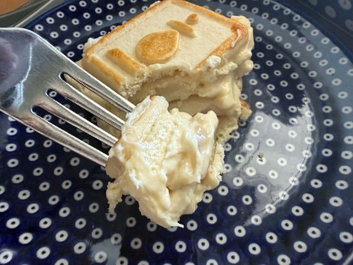 A slice of Chessmen banana pudding with a fork head held from the left with a bite on the tip of the fork sitting on a small, dark blue plate with white polka dots.