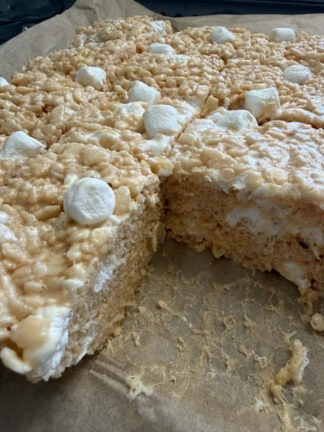 Squarely cut caramel rice crispy treats sitting on brown parchment paper with a piece missing from the right corner.