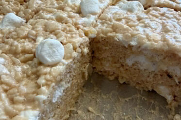 Squarely cut caramel rice crispy treats sitting on brown parchment paper with a piece missing from the right corner.