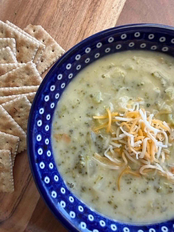 A dark blue Polish pottery bowl with white polka dots filled with Cream of Broccoli Soup and sprinkled cheese in the middle. To the left is a scattering of saltine crackers.