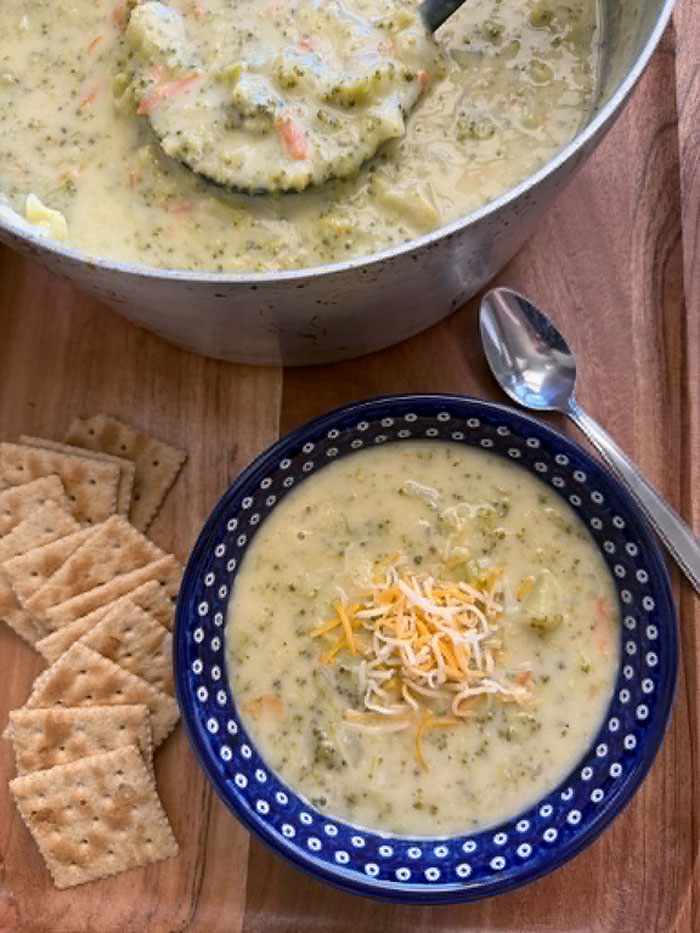 Cream of Broccoli Soup topped with shredded cheese in a dark blue bowl with white polka dots. To the left of the bowl are saltine crackers. A spoon is lying next to the bowl on the upper right. Above the bowl of soup is a partial picture of a pot of Cream of Broccoli Soup with a serving spoon head held directly above the soup with soup on it.