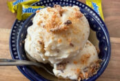 A small dark blue bowl with a couple of scoops of butterfinger ice cream topped with butterfinger bits with a spoon inserted from the left sitting on a small dark blue plate. A fun-size butterfinger candy bar is on the edge of the plate at the top, and two more butterfingers are lying near the plate at the top.