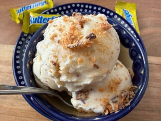 A small dark blue bowl with a couple of scoops of butterfinger ice cream topped with butterfinger bits with a spoon inserted from the left sitting on a small dark blue plate. A fun-size butterfinger candy bar is on the edge of the plate at the top, and two more butterfingers are lying near the plate at the top.