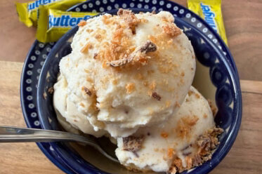 A small dark blue bowl with a couple of scoops of butterfinger ice cream topped with butterfinger bits with a spoon inserted from the left sitting on a small dark blue plate. A fun-size butterfinger candy bar is on the edge of the plate at the top, and two more butterfingers are lying near the plate at the top.