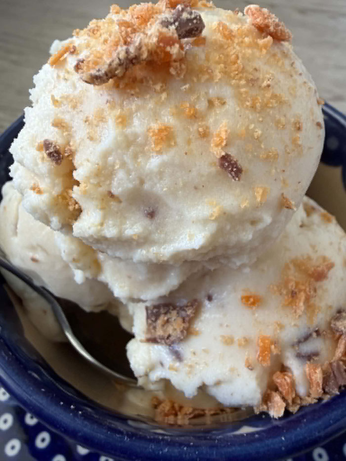 A close-up photo of three scoops of candy bar ice cream topped with crushed butterfingers in a small bowl with a spoon inserted from the left.