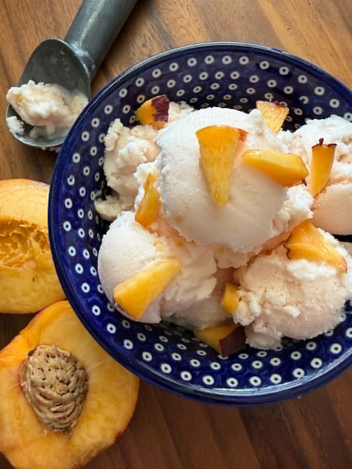 Several scoops of peach ice cream topped with small peach pieces in a dark blue and white polka-dotted bowl. On the left of the bowl are two peach halves. In the upper left corner is an ice cream scoop with a little bit of ice cream in it.