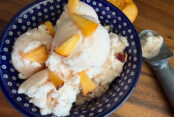 A dark blue and white polka-dotted bowl of peach ice cream with small slices of fresh peaches on top. On the right of the bowl is an ice cream scoop with a little bit of ice cream in it. A half a peach is near the right top of the bowl.
