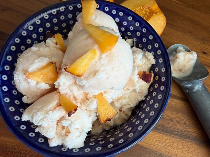 A dark blue and white polka-dotted bowl of peach ice cream with small slices of fresh peaches on top. On the right of the bowl is an ice cream scoop with a little bit of ice cream in it. A half a peach is near the right top of the bowl.