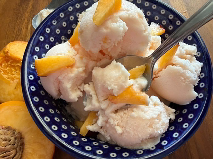 A dark blue and white polka-dotted Polish pottery bowl filled with peach ice cream and topped with small peach slices. A spoon is inserted diagonally from the right into the ice cream. On the left are two peach halves.