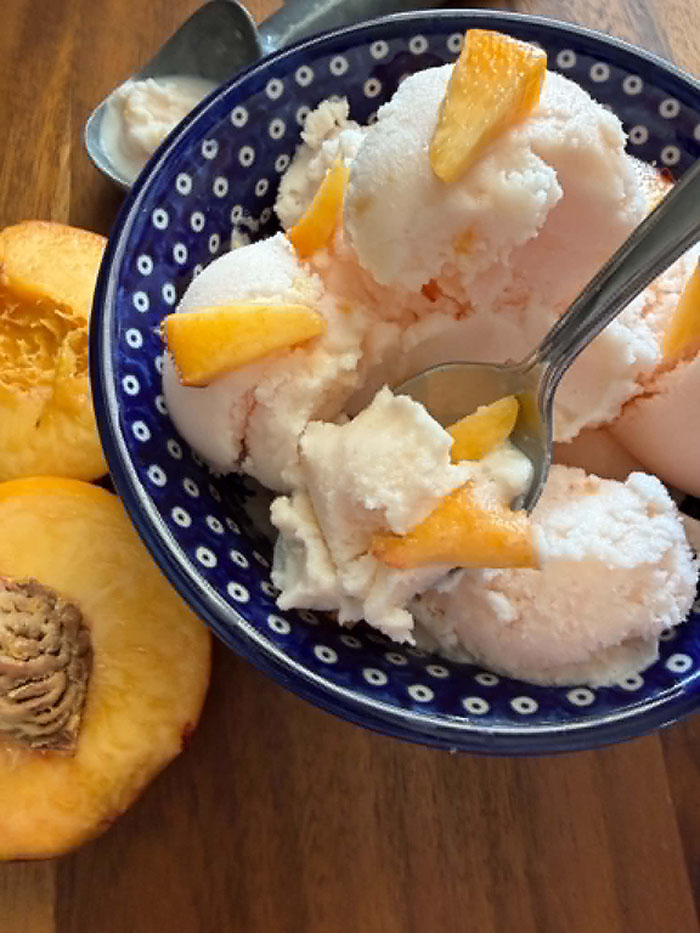 Two peach halves on the left of a dark blue white polka-dotted Polish pottery bowl filled with several scoops of peach ice cream topped with small fresh peach slices with a spoon inserted diagonally from the right into the ice cream. An ice cream scoop head is next to the upper right of the bowl.