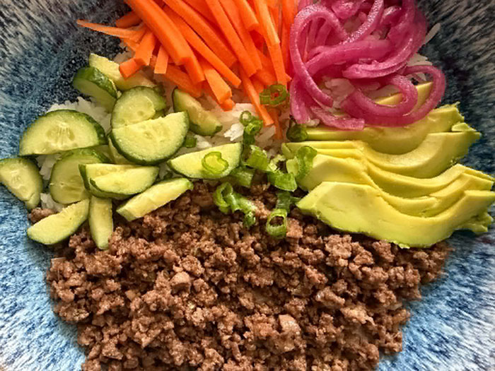 A bowl with quartered cucumbers, matchstick carrots, pickled red onion, sliced avocado, ground beef, and sliced green onion.