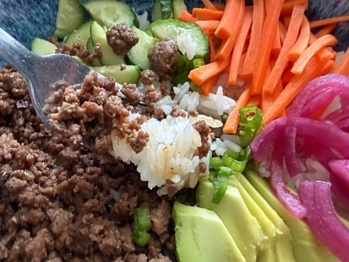 A fork head with ground beef and rice on top of a bowl of ground beef, sliced green onions, sliced avocado, pickled red onions, matchstick carrots, and quartered sliced cucumber.