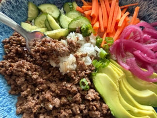 Seasoned ground beef, sliced cucumbers, sliced green onions, matchstick carrots, pickled red onions, and sliced avocado on rice with a fork held diagonally from the left with rice and ground beef on it directly over the ground beef.