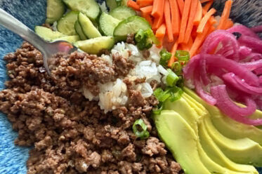 Seasoned ground beef, sliced cucumbers, sliced green onions, matchstick carrots, pickled red onions, and sliced avocado on rice with a fork held diagonally from the left with rice and ground beef on it directly over the ground beef.