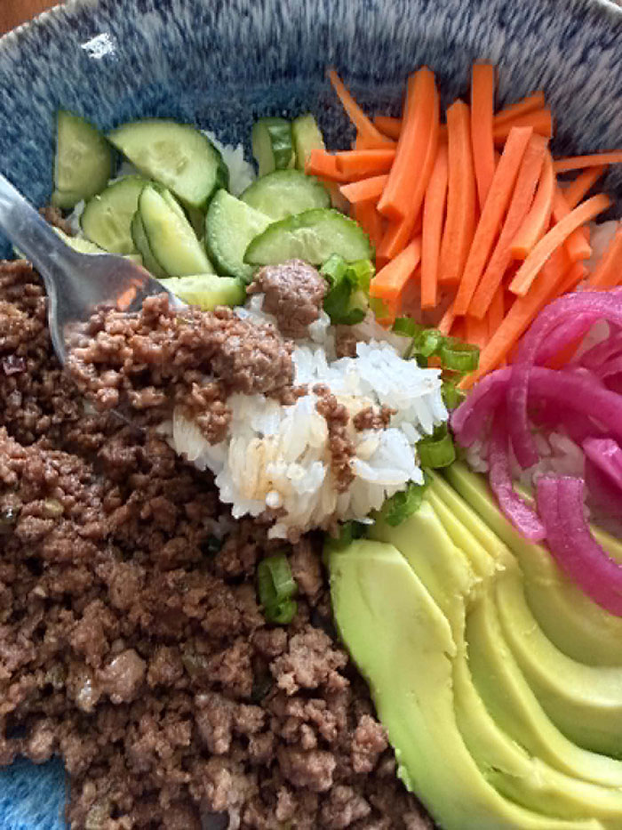 A closeup-photo of a bowl with rice on the bottom topped with sliced avocado, pickled red onions, matchstick carrots, sliced cucumbers, sliced green onions, and ground beef with a fork head with rice and ground beef lying diagonally from the left on top of the ground beef.