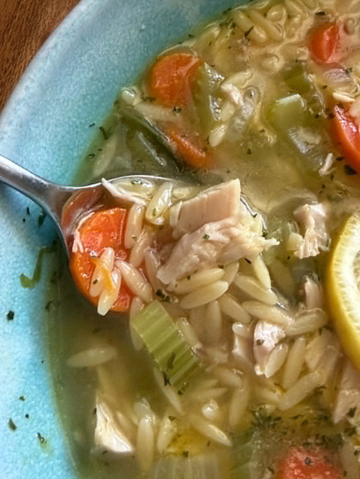 A partial picture of a bowl of Rotisserie Chicken Orzo Soup with a spoon lying diagonally on the edge of the bowl from the left with soup in it.