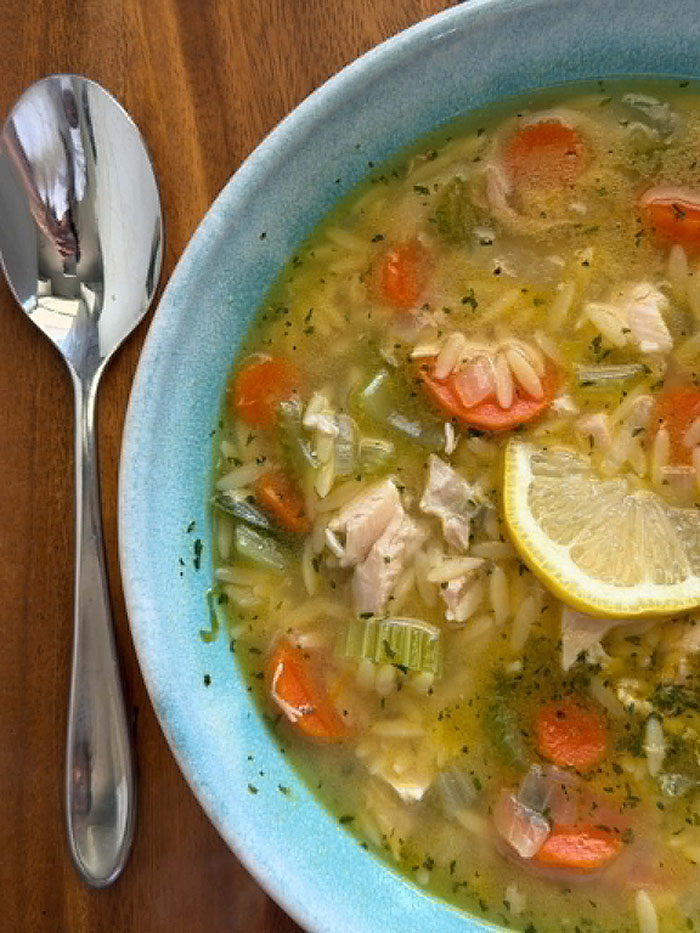 Rotisserie Chicken Orzo Soup in a blue bowl with a half slice of lemon in the middle and a spoon lying to the left of the bowl.