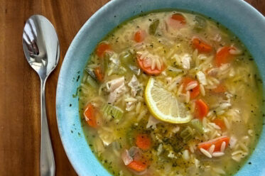 A bowl of Rotisserie Chicken Orzo Soup with a half lemon slice in the middle and a spoon on the left of the bowl.