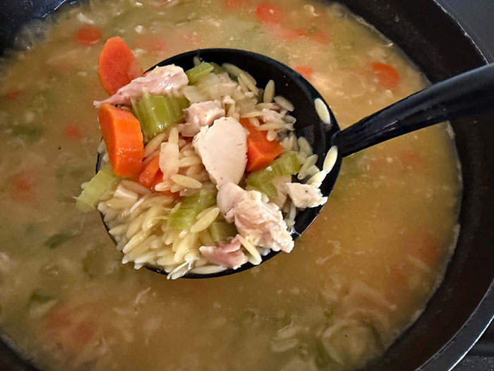 A pot full of Rotisserie Chicken Orzo Soup with a soup ladle held above from the right full of soup.
