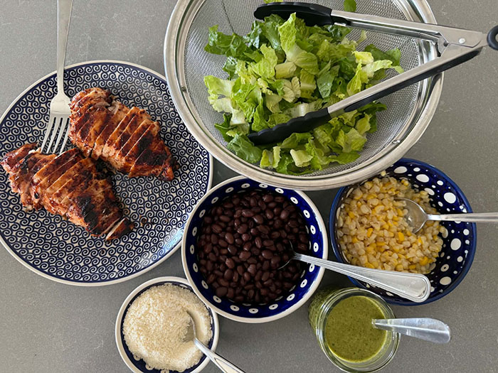 A plate with two sliced barbecued chicken breasts. A fork is inserted vertically into one of the chicken breasts. Next to the chicken breasts is a medium glass bowl with chopped romaine lettuce with tongs inserted from the right. A small bowl of corn with a spoon inserted from the right. A jar of cilantro lime vinaigrette with a spoon inserted into it. A small bowl of black beans with a spoon inserted from the right. A small bowl of Parmesan cheese with a spoon inserted.