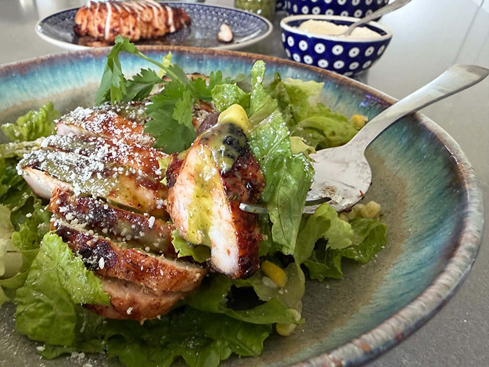 A dinner plate of BBQ chicken salad with cilantro lime vinaigrette with a fork lying on the plate and salad with a piece of chicken and romaine lettuce on it. In the background is a plate with sliced barbecue chicken breast and a small bowl of Parmesan cheese.