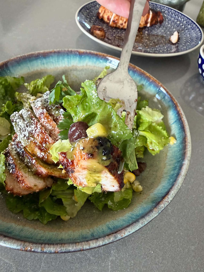 A plate of barbecue chicken salad with cilantro lime vinaigrette with a fork diagonally inserted in it. In the background is a plate with a sliced bbq chicken breast.
