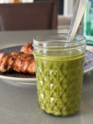 A small glass jar of cilantro lime vinaigrette with a spoon inserted and a plate of sliced barbecue chicken behind it.