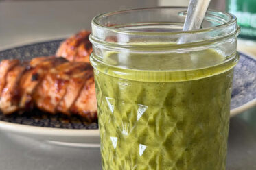 A small glass jar of cilantro lime vinaigrette with a spoon inserted and a plate of sliced barbecue chicken behind it.