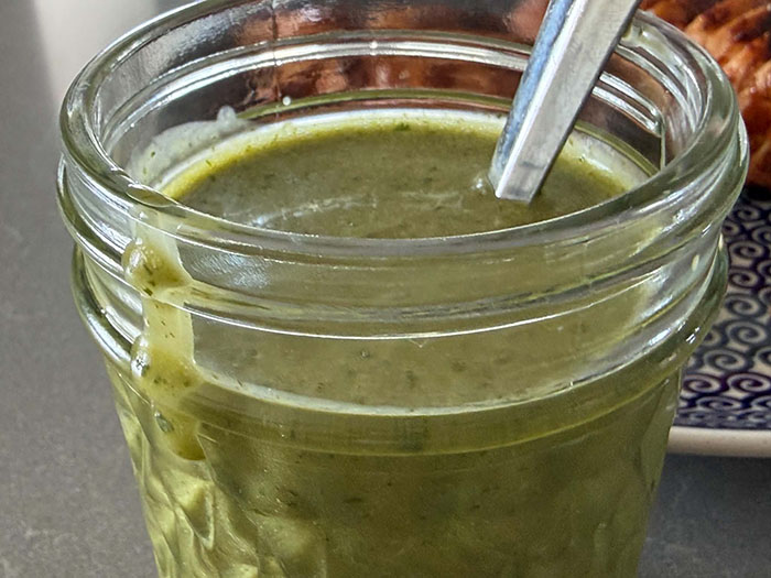 A close-up photo of a small glass jar of cilantro lime vinaigrette with a spoon inserted and some of the vinaigrette dripping down the side. 
