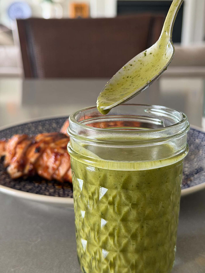A small glass jar of cilantro lime dressing with a spoon held diagonally above with dressing dripping off it. In the background is a plate with sliced barbecue chicken.