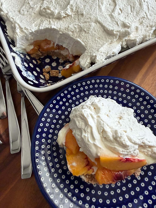 A serving of fresh peach dessert on a small dark blue plate with white polka dots. Next to the plate at the top is the corner of a 9x13 dish of Fresh Peach Dessert with a scoop taken out. Lying next to the dish and the plate are several forks.