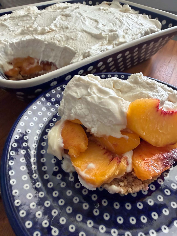 A 9x13 Polish pottery baking dish of peach dessert with a scoop taken out of the corner on the left. In front of the baking dish is a small dark blue plate with white polka dots with a scoop of fresh peach dessert.