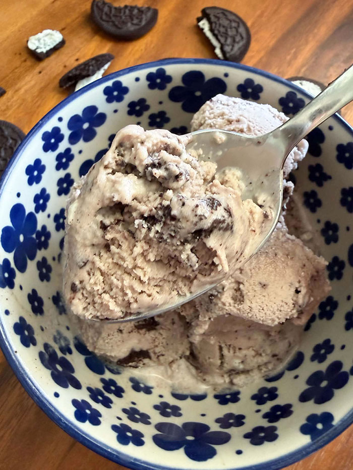 Homemade Oreo Ice Cream in a white bowl with dark blue flowers. A spoon is held above the bowl of ice cream with a scoop of ice cream on it. At the top, broken Oreos surround the bowl.