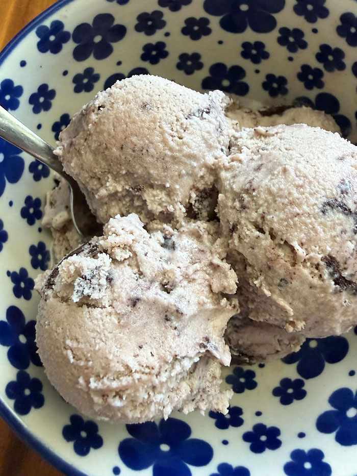 A white bowl with dark blue flowers with three scoops of Oreo ice cream. A spoon is inserted into the ice cream diagonally from the left.