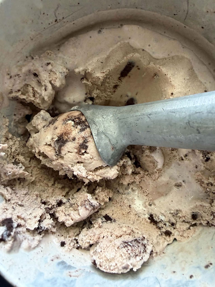 A metal canister of Homemade Oreo Ice Cream with an ice cream scoop with a scoop of ice cream lying diagonally from the right on top of the ice cream.