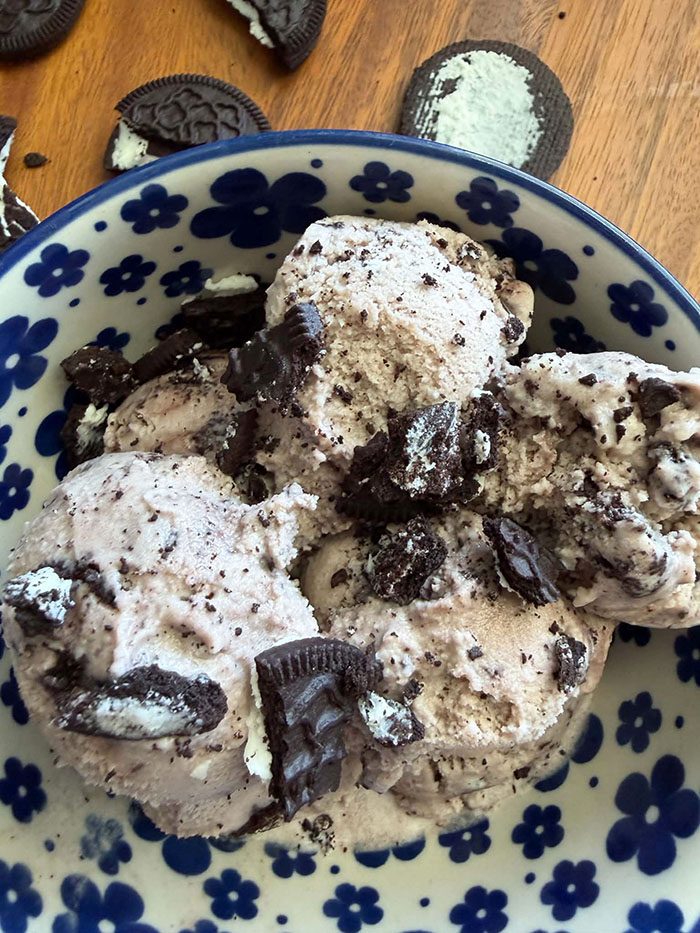 A white bowl with dark blue flowers with several scoops of Oreo ice cream with Oreo cookie pieces sprinkled on top. Several broken Oreo cookies surround the bowl at the top.