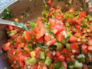 A metal mixing bowl of Jen's Pico de Gallo with a spoon with pico de gallo on it held from the left directly above.