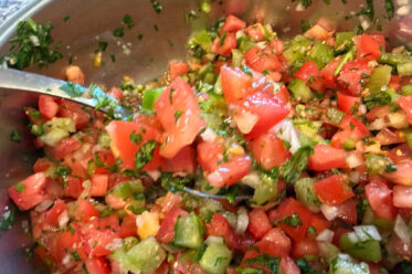 A metal mixing bowl of Jen's Pico de Gallo with a spoon with pico de gallo on it held from the left directly above.