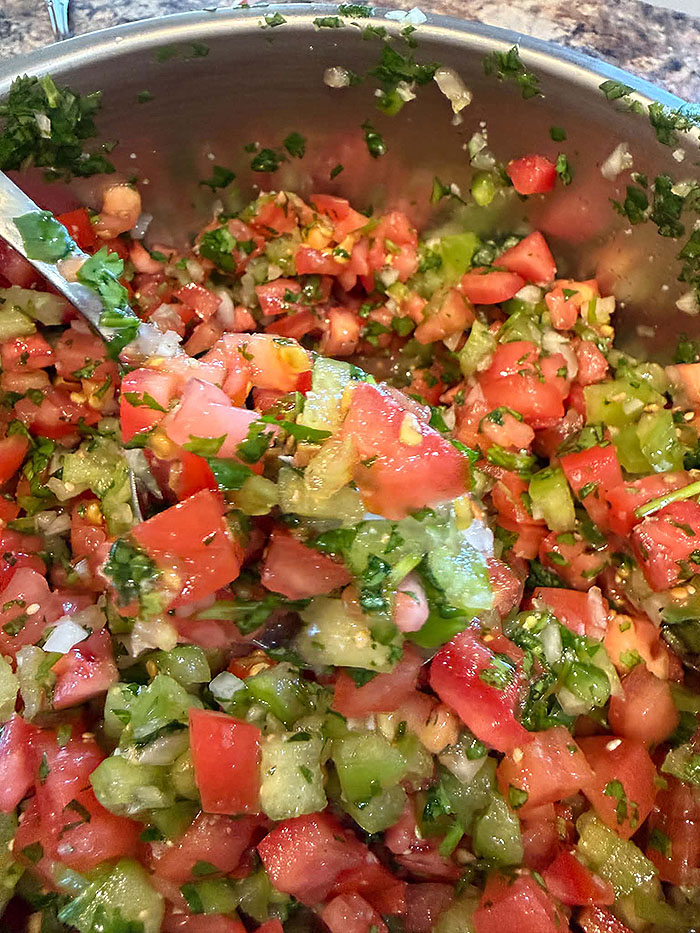 A medium-sized metal mixing bowl of pico de gallo with tomatillos. A spoon is diagonally resting on left side of the rim of the bowl with a scoop of pico de gallo on it.